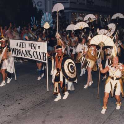Two people holding a Key West Woman's club banner with people walking behind them in the parade.