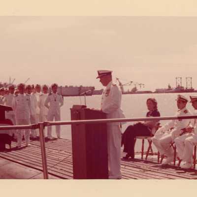 U.S. Navy men standing at attention and one speaking at the podium