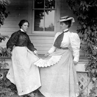 Deam05.jpg 534KB: Two women seated on a vine entwined porch in 1900s-style clothing. One woman is holding a fan