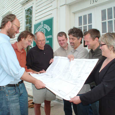 Posed publicity photo of planning team reviewing blueprints: (L to R) Nicolaas Wilkens, Steve Darpel, Tom Anthrop, Mike VandenBerg, Mark Schrock, Chuck Carlson, Amy Cook. (Wilkens, VandenBerg, Carlson and Cook represent architect/design group Fenn's Design Mill, Inc.; Darpel and Schrock are business partners in Darpel & Associates Builders Inc.; Anthrop is Old School House Task Force Chair)