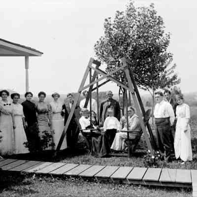 Sim Crow crowd.jpg 527KB: Presumed to be Crow family posed outdoors around a double-bench swing. The house is a farmhouse or cottage with a young tree and an empty field in the background.