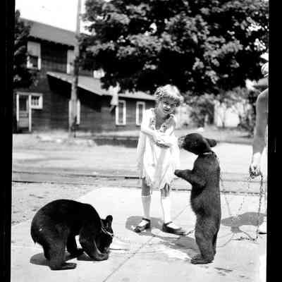 sim dancing bears mn129.jpg 802KB: A young girl bottle-feeds a chained bear cub while another cub sniffs the pavement. Behind the girl are railroad tracks and possibly the Saugatuck Fruit Exchange building.