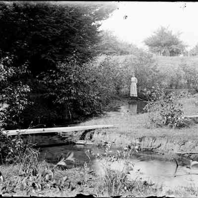 sim lady by stream.jpg 1.3MB: Woman standing along a stream with a plank bridge in the foreground.