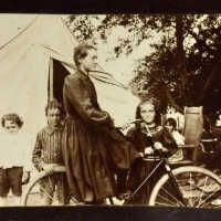          Melcher Family at Pier Cove, Michigan; Woman on bicycle in front of tent, with four children posing toward the camera.
   