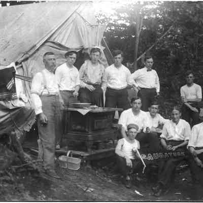 Campers_ca_1910.jpg 495KB: Men posed in their elaborate camp site that includes an iron stove and furniture. A young man is displaying a Saugatuck pennant.