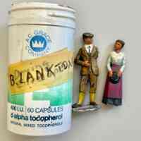          Two miniture human figurines made of painted, cast metal.; A man with wearing a cap and 3-piece suit with short pants and yellow socks. The man is missing his left arm. A woman in a white blouse and long dark skirt with purple apron, holding a hat. The woman is missing her base plate.
The figurines are in a plastic vitamin bottle with the a masking tape label that reads 