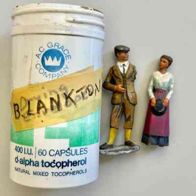Two miniture human figurines made of painted, cast metal.: A man with wearing a cap and 3-piece suit with short pants and yellow socks. The man is missing his left arm. A woman in a white blouse and long dark skirt with purple apron, holding a hat. The woman is missing her base plate.
The figurines are in a plastic vitamin bottle with the a masking tape label that reads "B. Lankton."