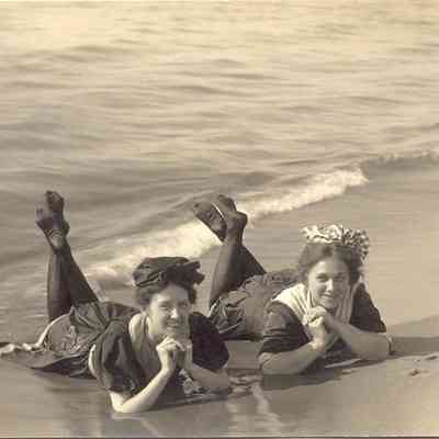 02.TwoLadiesLieonSaugBeach1910.jpg 101KB: Two women lay on the beach, in the shallows, their heads raised on their elbows and their ankles crossed. The two ladies are wearing dark swimming attire and stockings. The two girls are smiling.