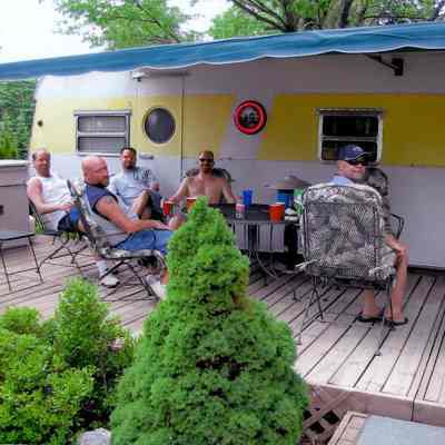 Camp_It2.jpg 718KB: Men enjoying the shade of a trailer's awning with deck furniture.