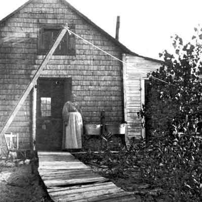Fishtownca1910tu.jpg 577KB - Digital file on Jack Sheridan Drive 2021.72.02: A mature woman stands beside a pair of washtubs outside a shanty's door