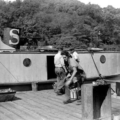 simmons-324-Fishing UnloadingFish: The tug Andrew was owned by Frank Sewers. It docked at Water Street, across the river from the Saugatuck-Douglas History Museum. A typical tug of the era, Andrew was enclosed to protect fishermen from the weather and to provide an enclosed workspace. Note buoy on roof of tug. Man dragging net boxes is Ev Van Plew