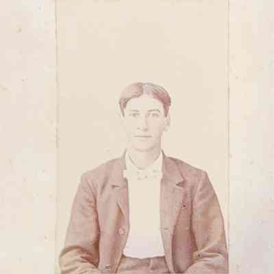 The image depicts a vintage portrait of a young man with neatly combed hair, dressed in a formal attire consisting of a dark suit and a bow tie, portrayed against a plain backdrop; the photograph, is framed in a simple border with some age-related discoloration and wear.