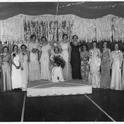 9 women standing on stage with four standing on a dais and one seated on a chair wearing a crown.