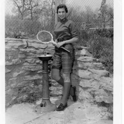 Student by water fountain and tennis court holding tennis racket.