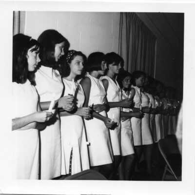 Students in white dresses holding candles inside a building.