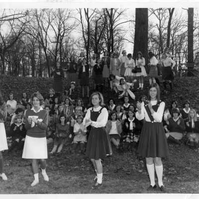 Four cheerleaders or club cheerleaders outside with student crowd behind them in 1964.