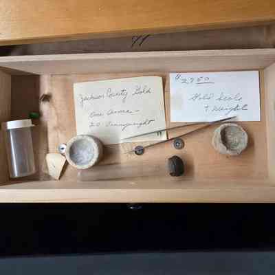 The image shows an open wooden drawer containing various small objects, including a clear cylindrical container, a sealed test tube, an old scale, two tiny stone bowls, and handwritten notes mentioning "Jackson County Gold" and prices, giving it an antique or collectible