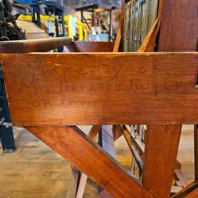 Caroline Stevens Rogers Loom, Inscription Detail.: The image shows a wooden structure with a handwritten inscription that reads "Property of Mrs. Horatio Rogers on loan." The wood appears to be part of a piece of machinery or furniture, possibly an antique loom, visible with its wooden frame and metal components. The background reveals a workshop or museum setting with various other mechanical parts and equipment visible. The lighting creates a warm, historical ambiance, suggesting an environment focused on preserving or displaying historical artifacts. The detailed craftsmanship of the wood and the aged appearance of the inscription add to the object's historical significance and authenticity.