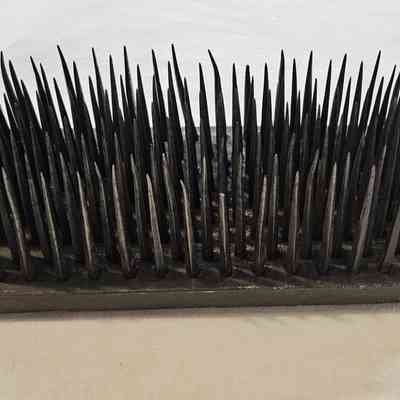 Hetchel: The image shows an old hetchel, a historical tool used for processing flax fibers. It consists of a wooden base with a rectangular shape, upon which numerous sharp, pointed metal spikes are arranged in dense rows. The spikes vary in length, creating a menacing appearance. Attached to the wooden base is a tag with some handwriting, likely catalog information for historical reference. This kind of tool was traditionally used to strip flax fibers from flax stems before spinning, playing an essential role in textile production. Its aged appearance suggests it has been well-used, with visible signs of wear and patina on both the wood and metal, reflecting its time-worn utility. The hetchel is displayed on a white or light-colored surface that contrasts with its dark tones, highlighting its intricate and spiky form.