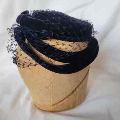 A vintage blue hat with netting is elegantly displayed on a wooden stand against a white background.