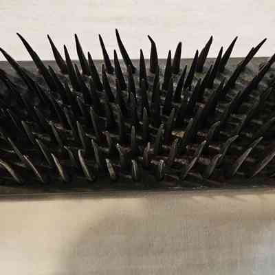 Hetchel, Top View: The image depicts a wooden base with an array of sharp, black, metallic spikes protruding upwards, resembling a dense field of thorns. The spikes are arranged in neat, parallel rows, each seemingly identical in shape and size, creating a menacing appearance. The base is rectangular, made of aged, textured wood with two metal brackets on either end, suggesting a secure attachment. A white string is tied to the base, attached to a small, rectangular tag with handwritten text, indicating possible archival or inventory details. The overall appearance of the object is archaic and utilitarian, possibly a tool or device from a bygone era, lying on a plain, neutral-colored surface that contrasts with the dark spikes, highlighting their stark, ominous presence.