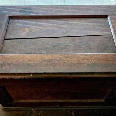Sewing Machine Lid/Cover. After Cleaning.: The image depicts an old, rectangular wooden box with a flat lid and a slightly raised border around the top edge, showing signs of wear and age with a visible crack on its surface, set against the backdrop of partially open blinds that allow natural light to illuminate the box, highlighting its worn and rustic appearance; the warm, brown tones of the wood convey a sense of history and character.