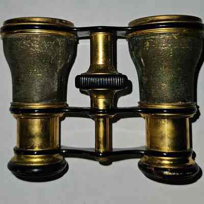 Opera Glasses, Top View: The image depicts a pair of vintage brass opera glasses with a well-worn patina, indicating age and frequent use. The eyepieces are set in a framework that includes a central focusing mechanism with a textured black knob, enabling the adjustment of the lenses for clear viewing. The glasses have a classic, symmetrical design, with smooth curves and dark accents that add to their antique aesthetic. The sheen of the brass contrasts with the darker elements, highlighting the craftsmanship of this elegant, historical optical instrument, which rests on a plain white background, emphasizing its intricate details.