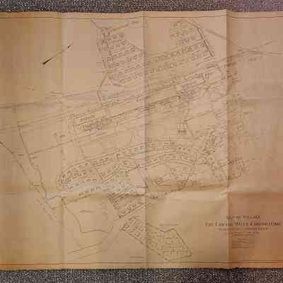 Map: The image shows a detailed map titled "Map of Village, The Lawton Mills Corporation, Plainfield, Connecticut," dated 1921. The map features a layout of the village, showcasing a network of roads, plots, and buildings. The central area is dominated by the Lawton Mills factory, surrounded by residential streets lined with small houses. Each house is marked by a rectangular shape, while the roads are labeled with names, providing a sense of the village's organization. The map includes small icons and symbols representing different landmarks or features, such as mills and other structures, and is drawn on aged paper, giving it a historical and intricate appearance. The map is oriented with a north arrow at the top left and bears a scale indicating distances.