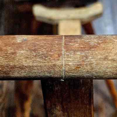 Yarn Winder, Take-up Bracket on End of Arm.: The image displays a close-up view of a wooden take-up bracket, possibly part of a tool or piece of furniture, featuring a cylindrical shape with round flanges on each end and a textured, grainy surface typical of natural wood. The handle extends horizontally across the image, occupying most of the foreground, while the background fades into a blurred wooden texture, suggesting a rustic or natural setting that enhances the focus on the craftsmanship of the handle. The perspective and lighting highlight the subtle variations and imperfections in the wood, lending an authentic and earthy aspect to the overall composition.