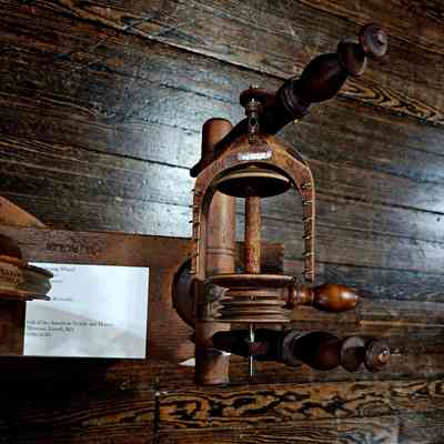 Flyer of Spinning Wheel: The image depicts a vintage spinning wheel set against a wooden floor backdrop, showcasing intricate wooden craftsmanship with multiple handles and a central spindle. The well-preserved antique, likely made in the 19th century, features detailed woodwork typical of historical textile machinery, reflecting the industrial heritage of the era. A small sign attached to the structure provides information, indicating that this spinning wheel is part of a collection contributed by the American Textile and History Museum located in Lowell, Massachusetts, inventory number 1890.16.83. The warm brown tones of the wood and the visible grain patterns emphasize the wheel’s historical and aesthetic appeal, making it an intriguing relic of past textile production methods.