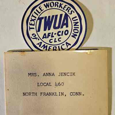 Conference Attendee Badge: The image displays a badge and a card. The badge, with a circular design, reads "TWUA AFL-CIO CLC, Textile Workers Union of America," in bold blue letters, reflecting an association with a labor union. Below the badge, there's a card with typed text that says "Mrs. Anna Jencik, Local 460, North Franklin, Conn.," indicating membership details for an individual associated with a local union chapter. The items shown suggest a historical or archival context related to labor or trade unions in America.