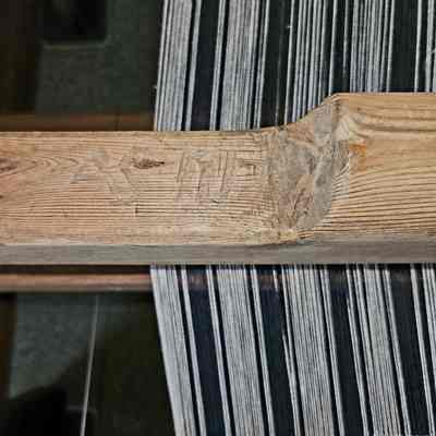 Maker's initials and date of construction carved into the beater bar.: The image shows a close-up view of an intricately woven textile on a wooden loom. The loom's wood has a rustic appearance, possibly indicating significant use over time, with the letters "AS" and date "1717" etched into the beater bar. The fabric being crafted features parallel black and white threads, suggesting a piece of cloth in the process of being woven. The photograph captures the detailed craftsmanship and the traditional art of weaving, highlighting the texture of both the wood and the fabric, with light subtly casting shadows to enhance the sense of depth and texture in the materials.