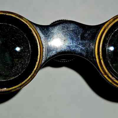Opera Glasses, Front View: The image shows a pair of vintage opera glasses viewed from the front, highlighting the large, round lenses surrounded by a well-worn brass or metal casing. The binoculars have a slightly aged appearance with some visible scratches and minor corrosion on the surface, suggesting they have been used extensively over time. The reflective glass lenses catch the light, adding a bright contrast against the darker material of the casing. Positioned against a plain, light-colored background, the binoculars are presented as the central focus of the image, showcasing their historical and functional design. The binoculars appear to be crafted from durable materials that offer a glimpse into an earlier era of optical equipment, reflecting both the artistry and engineering of their time. The overall composition captures the blend of functionality and beauty inherent