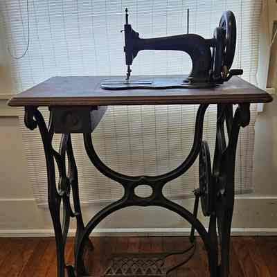 Sewing Machine with Lid/Cover Removed. Front View. Before Cleaning.: The image shows an antique sewing machine with a cast iron base and a wooden tabletop, featuring a traditional design typical of older, manually-operated sewing machines. The machine stands prominently against a backdrop of soft light filtering through a window with blinds, casting subtle shadows on the hardwood floor. The intricate metalwork includes ornate, curved designs that serve as both functional and decorative elements. The foot pedal is visible beneath the table, indicating its use for manual operation, and the overall appearance suggests a piece that blends historical craftsmanship with practical functionality, likely evoking a sense of nostalgia.