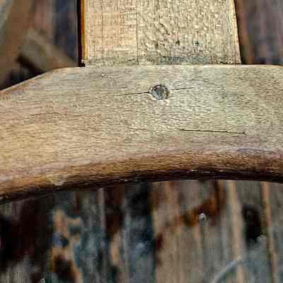 Yarn Winder, Take-up Bracket on End of Arm.: The image shows a close-up of a rustic, wooden take-up bracket attached to a rectangular piece of wood, possibly part of a tool or furniture item. The bracket has a smooth, curved design and an aged appearance with visible grain and small cracks, suggesting it has been used over time. The background is composed of dark, wooden planks with a weathered texture, adding to the overall vintage and natural aesthetic of the composition. The lighting enhances the wood's warm tones, highlighting its natural imperfections and character.