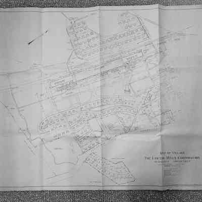 Map, Black and White Photo: The image depicts a detailed map of a village created by The Lawton Mills Corporation in Plainfield, Connecticut. The map outlines various plots of land, buildings, roads, and natural features within the village. It includes labeled sections for residential areas, industrial buildings, and public spaces, along with pathways and waterways. Key landmarks and geographical markers are noted, and the map is scaled, with notations and measurements to guide the reader. A compass rose is present, indicating directions.