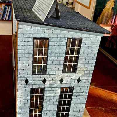 Right Side View: The image shows a detailed miniature model of a gray brick building with multiple windows and a shingled roof, displaying a sign on top. Inside, the model appears to depict interior scenes visible through the windows. The building is set on a wooden base, placed on a wooden table.