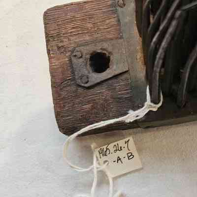 Hetchel: The image shows a close-up of a rustic wooden object with an aged, dark finish and a textured surface, featuring a metal plate with a hole in the center attached to it, secured with visible screws. To the right, part of a metal structure is visible, possibly composed of rods, indicating some form of historical or industrial use. A piece of white string is attached to the metal component, with a handwritten label beneath it, detailing the numbers "1965.26.7 2-A-B," suggesting the item might be part of a collection or archival system. The whole setup is placed on a light-colored fabric background, giving the scene an organized and archival appearance. The combination of materials and labeling suggests this could be a museum artifact.