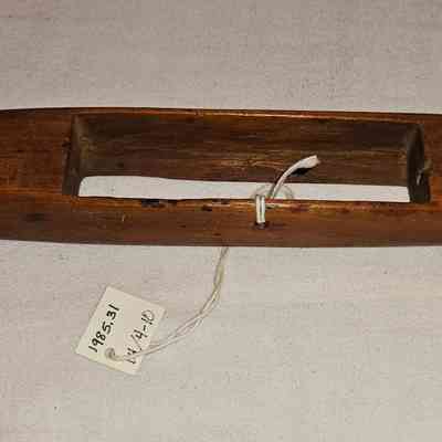Throw Shuttle, Bottom: The image displays a wooden boat shuttle, which is an elongated, smooth tool with tapered ends and a hollow center, traditionally used in weaving to carry the weft thread back and forth across the warp on a loom. It features a small, tied white string with a tag attached, on which numbers and possibly identifiers are written, indicating perhaps an inventory or catalog number. The shuttle rests on a plain, light-colored fabric surface that provides a neutral background, highlighting the warm, natural tones of the wood, along with its aged, rustic appearance, which suggests it has been used over the years in a weaving or textile setting, perhaps for demonstration or educational purpose in crafting.