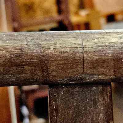 Yarn Winder, Take-up Bracket. Outer flange is missing, to make it easier to slide the completed skein off the winder.: The image shows a close-up view of the top part of a vintage wooden take-up bracket. The wood has a rich, aged texture with visible grains and a few nicks, suggesting considerable use over time. The background is blurred, containing other wooden furniture with various shapes, hinting at a setting like a workshop or antique store. The lighting is warm and natural, enhancing the rustic ambiance of the scene and emphasizing the character and history embedded in the worn, smooth finish of the wood.