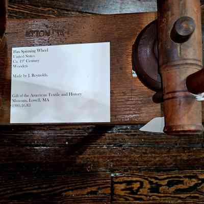 Spinning wheel showing inscription "J. Reynolds": The image shows a close-up of a flax spinning wheel made in the United States during the 19th century. Constructed from wood, this historical artifact was crafted by J. Reynolds. Accompanying the spinning wheel is a label indicating that it is a gift from the American Textile and History Museum located in Lowell, Massachusetts, with the catalog number 1980.16.83. The label is placed on a wooden surface, which bears visible wood grain and subtle signs of aging, enhancing the vintage appearance of the spinning wheel. The artifact is an example of textile production tools from its era.