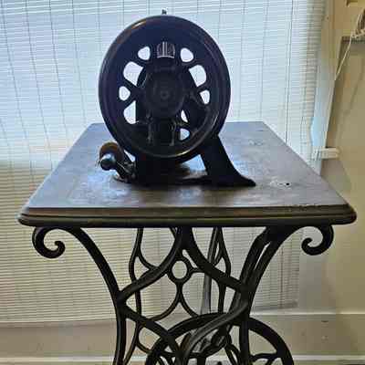 Sewing Machine. End View. Before Cleaning.: The image shows an antique sewing machine with an intricately designed metal base, featuring ornate scrollwork and a large flywheel. The machine sits atop a wooden tabletop with an aged, rustic appearance, positioned against a backdrop of vertical blinds that allow diffused light to filter into the room. The hardwood floor beneath the sewing machine adds to the vintage aesthetic, complementing the historical charm of the piece. The craftsmanship of the metalwork is both functional and decorative, illustrating the design sensibilities of a bygone era when such devices were not just tools but also integral parts of home decor.