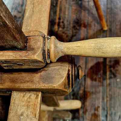 Yarn Winder, Crank Handle.: The image shows a close-up view of a traditional wooden tool featuring a handle attached to a mortise and tenon joint, possibly part of a larger woodworking device or furniture piece. The wood appears aged with visible grain patterns and a patina that suggests frequent use over time. The handle is cylindrical and smoothly polished, indicating it was well-crafted for ergonomic grip. Below the joint, the wooden surface is slightly worn, further emphasizing the item's age and usage history. The background reveals more wood, adding context to the authentic, rustic setting.