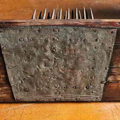 Hetchel, bottom view: The image shows a rustic, antique-looking wooden tool, a flax comb or hetchel, with a row of sharp, metal spikes protruding from the top side. The wood appears weathered, with a rich, dark brown color and visible grain patterns, indicating age and frequent use. A strip of heavily rusted metal is affixed to the bottom surface of the wooden structure, secured by numerous unevenly spaced nails, reflecting a utilitarian yet worn-out appearance. The background is a wooden surface with a similar texture but a lighter hue, creating a harmonious yet rustic atmosphere that suggests an old workshop setting.