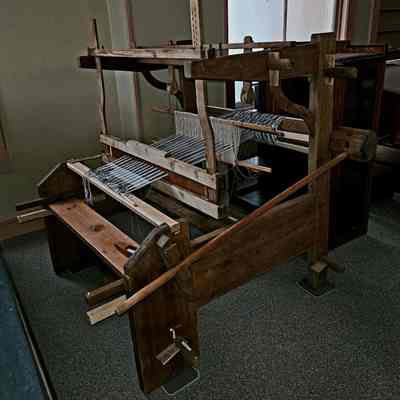 Fully assembled and conserved -- in working order.: The image depicts a traditional wooden loom, positioned in a dimly lit room with neutral-colored walls. This loom, made from sturdy wood with a natural finish, features horizontal beams and numerous strings of warp thread lined up meticulously. It appears to be quite old and well-crafted, showcasing a classic design with wooden pegs and paddles. The device occupies a significant space on the carpeted floor, reflecting the historical artisan craftsmanship used for weaving textiles. Light from a window softly illuminates the scene, casting gentle shadows that emphasize the loom’s intricate structural details.