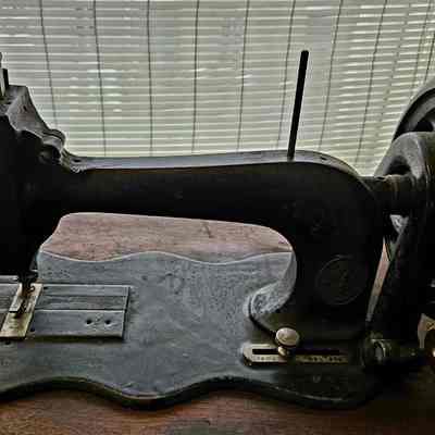 Close Up Photo of the Sewing Machine. Before Cleaning.: The image depicts an antique sewing machine, featuring a dark, heavy metal frame with intricate detailing and a large, spoked flywheel on the right side, resting on a wooden surface. The machine's design showcases the craftsmanship typical of early sewing devices, with a metal arm extending horizontally over a flat base that includes a needle and presser foot area. The background consists of a window covered with white blinds, allowing soft, diffused light to illuminate the scene, highlighting the sewing machine’s vintage charm and well-worn patina, suggesting its historical significance and the stories it might hold from generations past.