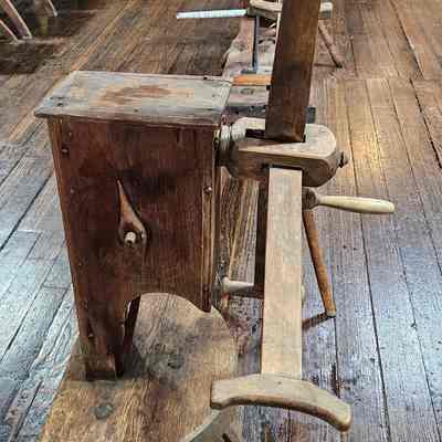 Yarn Winder, Side View.: The image depicts a vintage wooden device known as a yarn winder of click reel. The wood is aged and has a weathered appearance, reflecting its historical use. This device is positioned on a polished wooden floor within a room that seems to be furnished with wooden chairs and structural elements. The lighting highlights the texture of the wood and the craftsmanship involved. Overall, the image evokes a sense of historical practices and the construction techniques of past eras.
