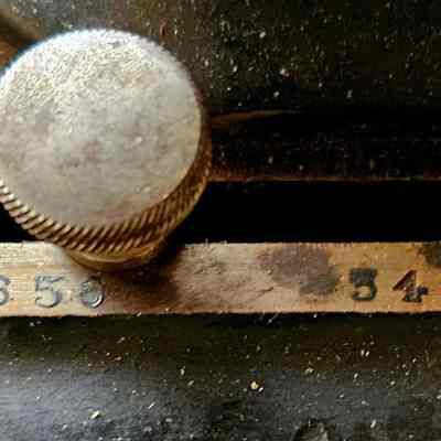 Sewing Machine, Close Up of Serial Numbers. Before Cleaning.: The image shows a close-up view of a mechanism featuring a metal knob with a textured grip positioned above a slot, adjacent to a sequence of embossed numbers: "440658" and "341656." The surface surrounding the knob appears to be aged and slightly worn, suggesting that the device or equipment is vintage or has been extensively used. The numbers are printed in a contrasting color for visibility against the darker background, and the surrounding area shows signs of wear, with some dust and a few spots of discoloration, indicating a patina developed over time.