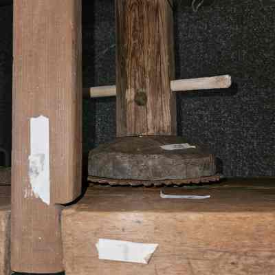 Iron or steel gear.: The image depicts a close-up view of a rustic wooden loom featuring a horizontal beam with a noticeable textured surface  and small cylindrical wooden supports extending horizontally on either side. The end of the horizontal beam intersects a vertical beam. A metal disc, which appears slightly corroded, is anchored to both pieces of wood. The surrounding area shows a dark, textured background that enhances the aged appearance of the wood. Several pieces of tape are affixed to the wooden components, serving as markers or indicators, adding to the work-in-progress or maintenance aspect suggested by the setup. Overall, the composition suggests a constructed assemblage, with visible signs of wear and repair.