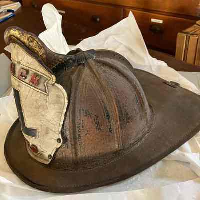 Side view of fire helmet: This image shows a view of the helmet from the right side.  It is made primarily out of brown leather with white leather in the front which extends from the cap. The white portion appears to be bolted on. In the center of the cap on the top is a golden metal eagle which latches onto the top of the white portion. There is some fading of the brown leather on the sides showing some black underneath. There is a clip on the back of the helmet.